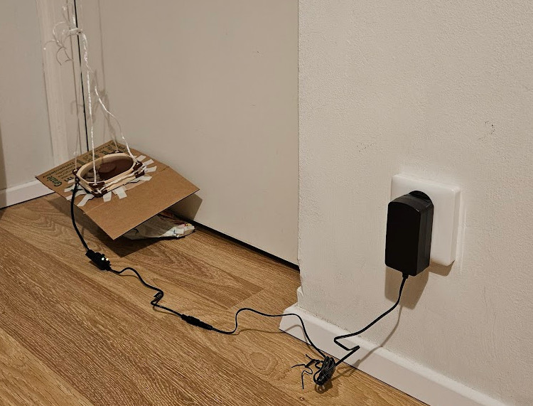 A photo of the prototype made from pizza boxes and tape, suspended near the floor by the door.